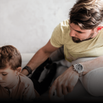 A father and son are lying on a white couch together, both holding video game controllers. The father, wearing a light yellow t-shirt and a silver watch, watches his son closely. The young boy, focused and engaged, wears a light pink shirt. The background features soft natural light and a neutral-toned wall.