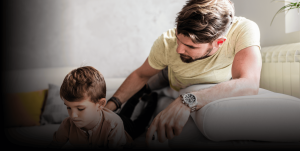 A father and son are lying on a white couch together, both holding video game controllers. The father, wearing a light yellow t-shirt and a silver watch, watches his son closely. The young boy, focused and engaged, wears a light pink shirt. The background features soft natural light and a neutral-toned wall.
